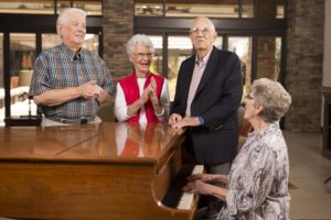 group of older adult men and women singing and clapping around woman playing a grand piano