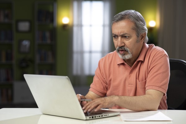 An older man with gray hair and a beard sits at a desk, looking intently at a laptop screen with a concerned expression. He's wearing a salmon-colored polo shirt and appears to be in a home office or study, with bookshelves visible in the background.